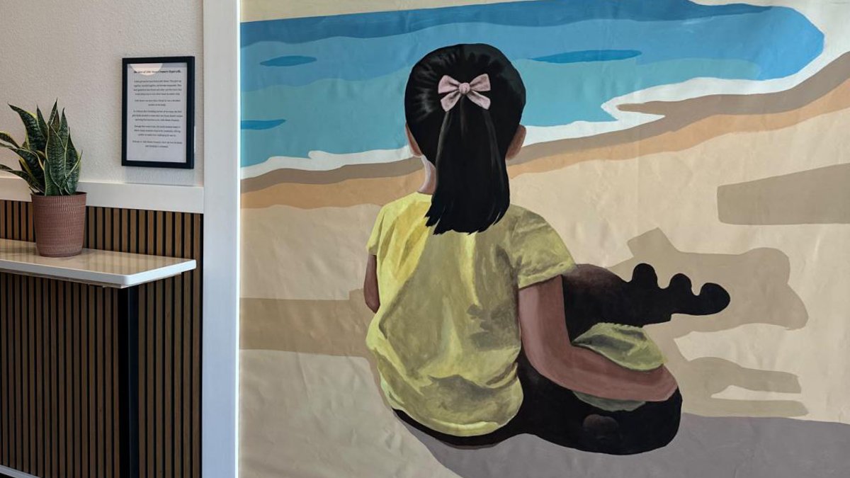 Interior feature wall mural at Little Moose Creamery in Frisco, Texas, depicting the owners' young daughter from behind wearing a yellow shirt and pink bow, sitting on a sandy beach next to her moose plush toy.