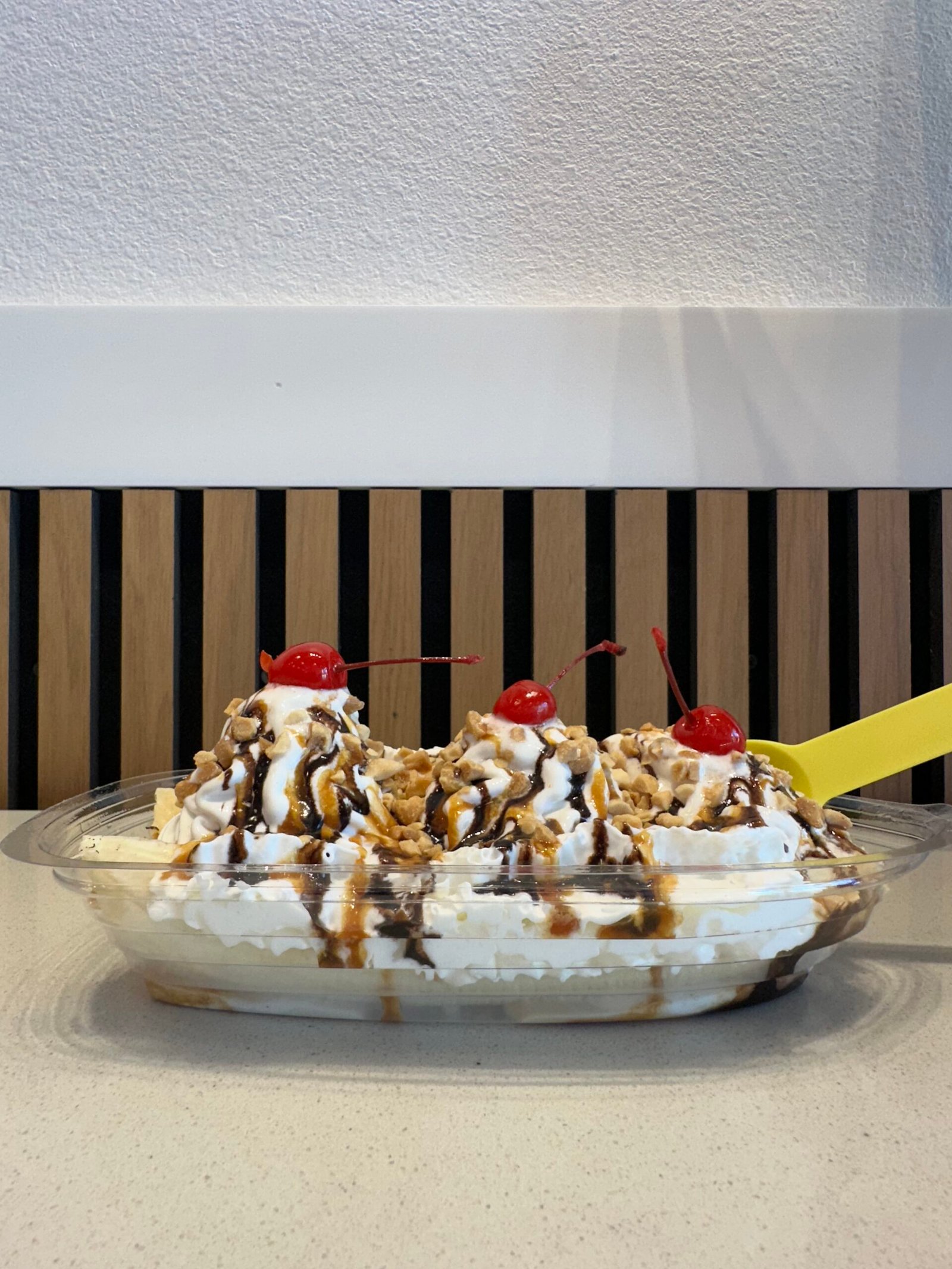 A Campfire Banana Split from Little Moose Creamery in Frisco, TX, soft serve ice cream topped with whipped cream, fresh banana, crushed peanuts, chocolate & salted caramel sauces, and cherries in a clear dish on the counter.
