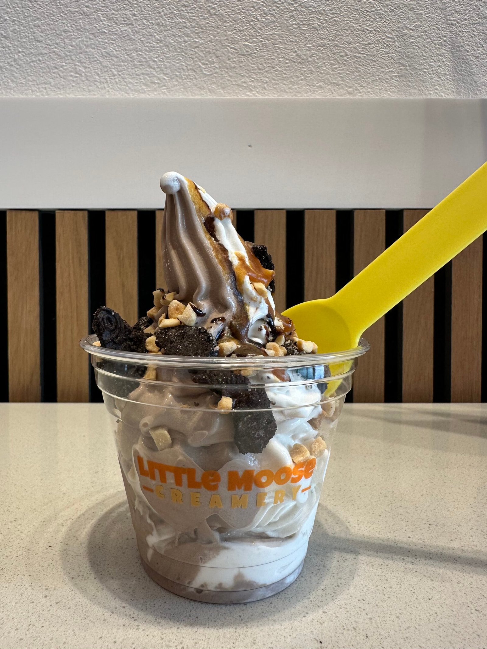 Kitchen Sink Sundae in a clear cup with the Little Moose Creamery logo, featuring layered chocolate and vanilla soft serve, topped with crushed cookies, chopped nuts, drizzle, and a yellow spoon.