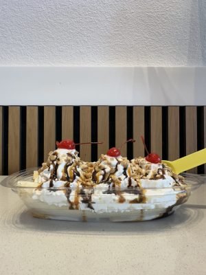 A Campfire Banana Split from Little Moose Creamery in Frisco, TX, soft serve ice cream topped with whipped cream, fresh banana, crushed peanuts, chocolate & salted caramel sauces, and cherries in a clear dish on the counter.