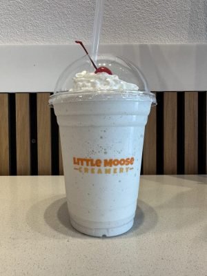 A branded clear plastic cup from Little Moose Creamery filled with a white vanilla milkshake, topped with swirled whipped cream and a cherry.