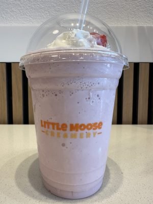 A bright pink strawberry milkshake in a clear cup with the Little Moose Creamery logo, capped with whipped cream and a cherry on top.
