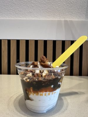 A Turtle Sundae from Little Moose Creamery in a clear plastic cup, featuring vanilla soft serve layered with rich chocolate sauce, salted caramel, and topped with whole crunchy pecans and a bright yellow spoon.